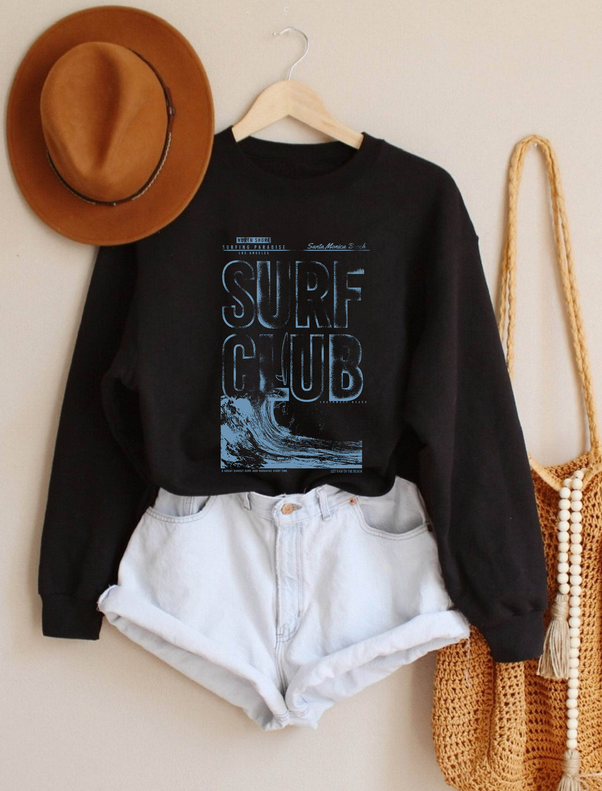 A black crewneck sweatshirt with a blue 'SURF CLUB' graphic print, styled on a wooden hanger with white denim shorts, a brown wide-brim hat, and a woven straw bag against a beige wall.