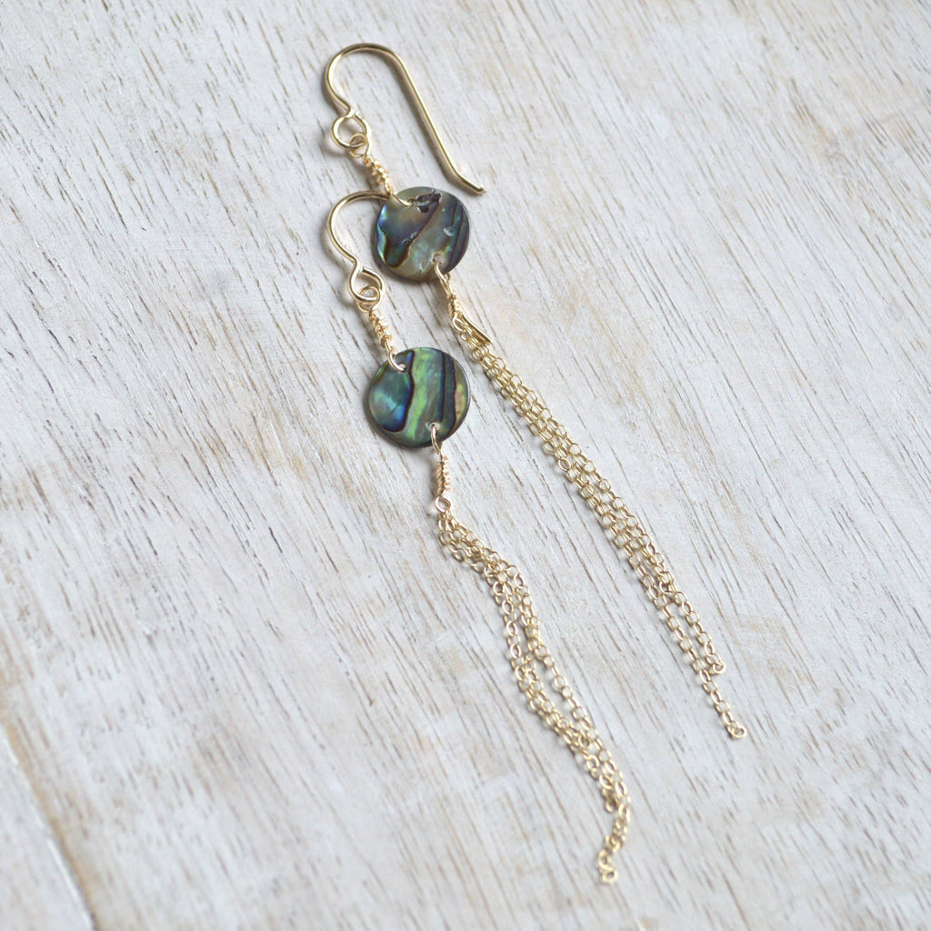 A pair of gold dangle earrings with round iridescent abalone shell beads and long delicate gold chains, displayed on a light-colored wooden surface.