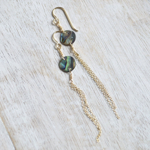 A pair of gold dangle earrings with round iridescent abalone shell beads and long delicate gold chains, displayed on a light-colored wooden surface.