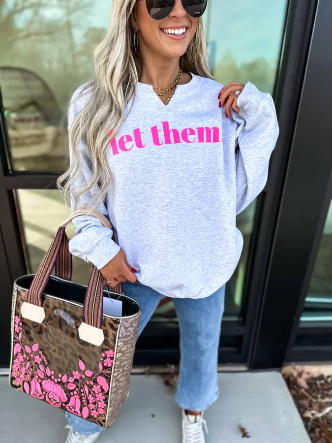 Woman wearing a white sweatshirt with pink text, holding a floral handbag, standing outside.