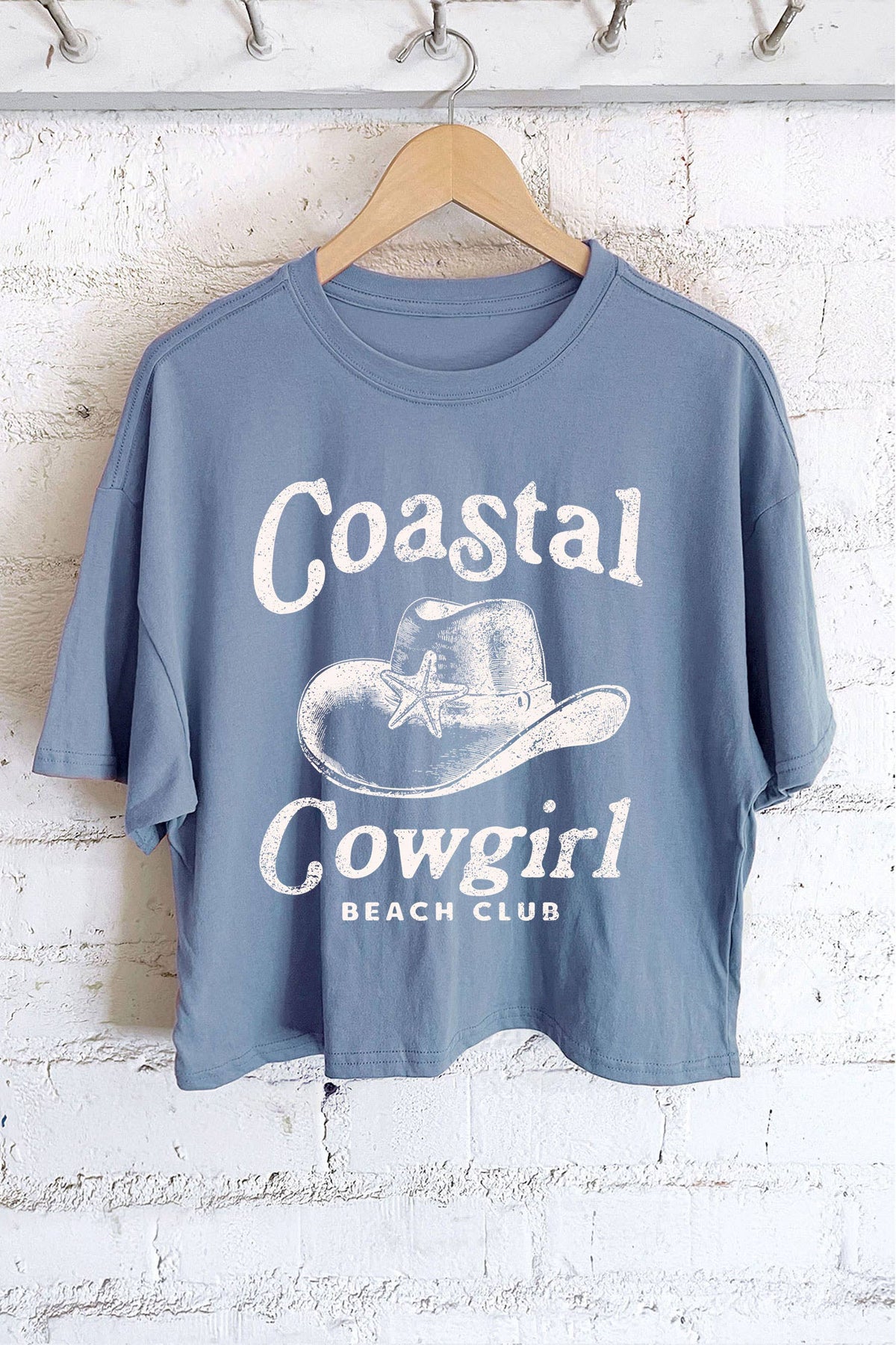 Blue t-shirt with 'Coastal Cowgirl Beach Club' text and graphic on a hanger against a white brick wall.