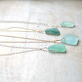 Three sea glass pendants in light aqua green, each attached to a delicate chain necklace in silver and gold tones, arranged on a light wood background.