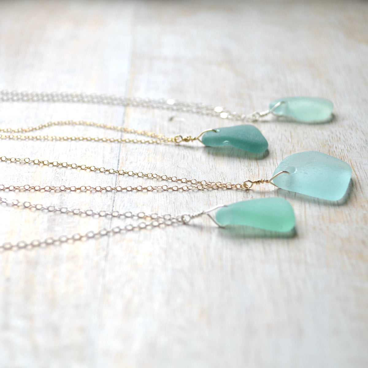 Three sea glass pendants in light aqua green, each attached to a delicate chain necklace in silver and gold tones, arranged on a light wood background.