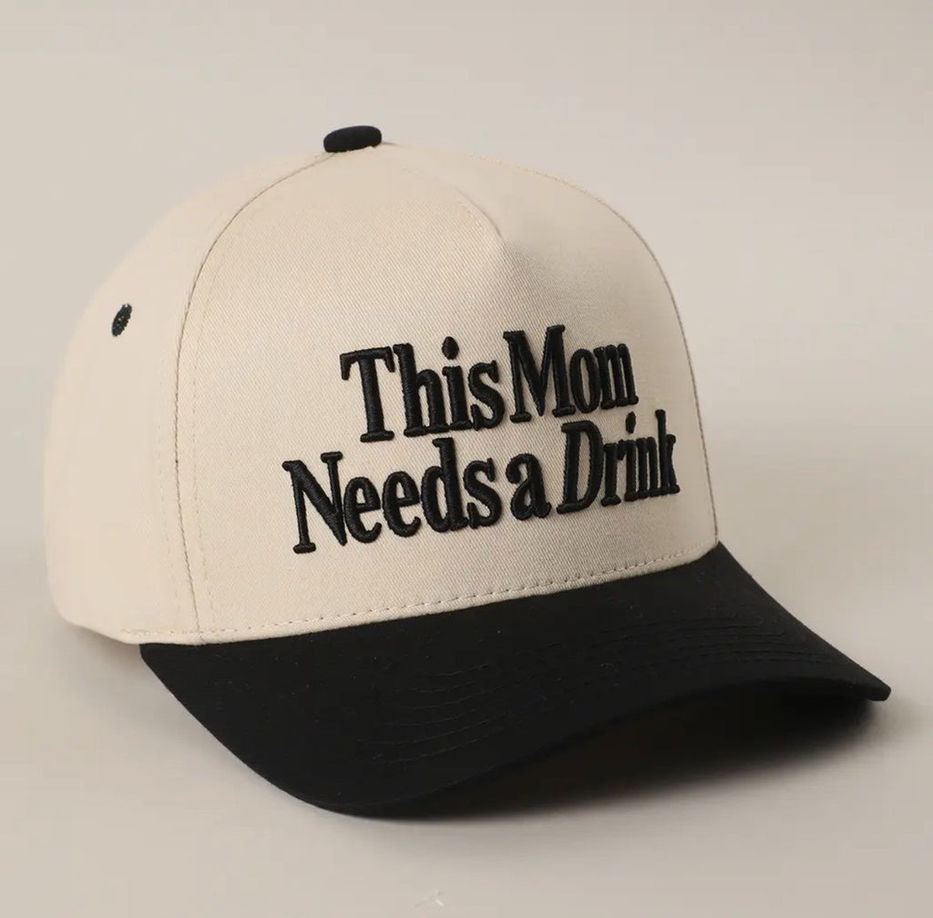 A baseball cap with an off-white crown and a black brim, featuring the text 'This Mom Needs a Drink' embroidered in black on the front. The cap is photographed on a neutral light gray background.