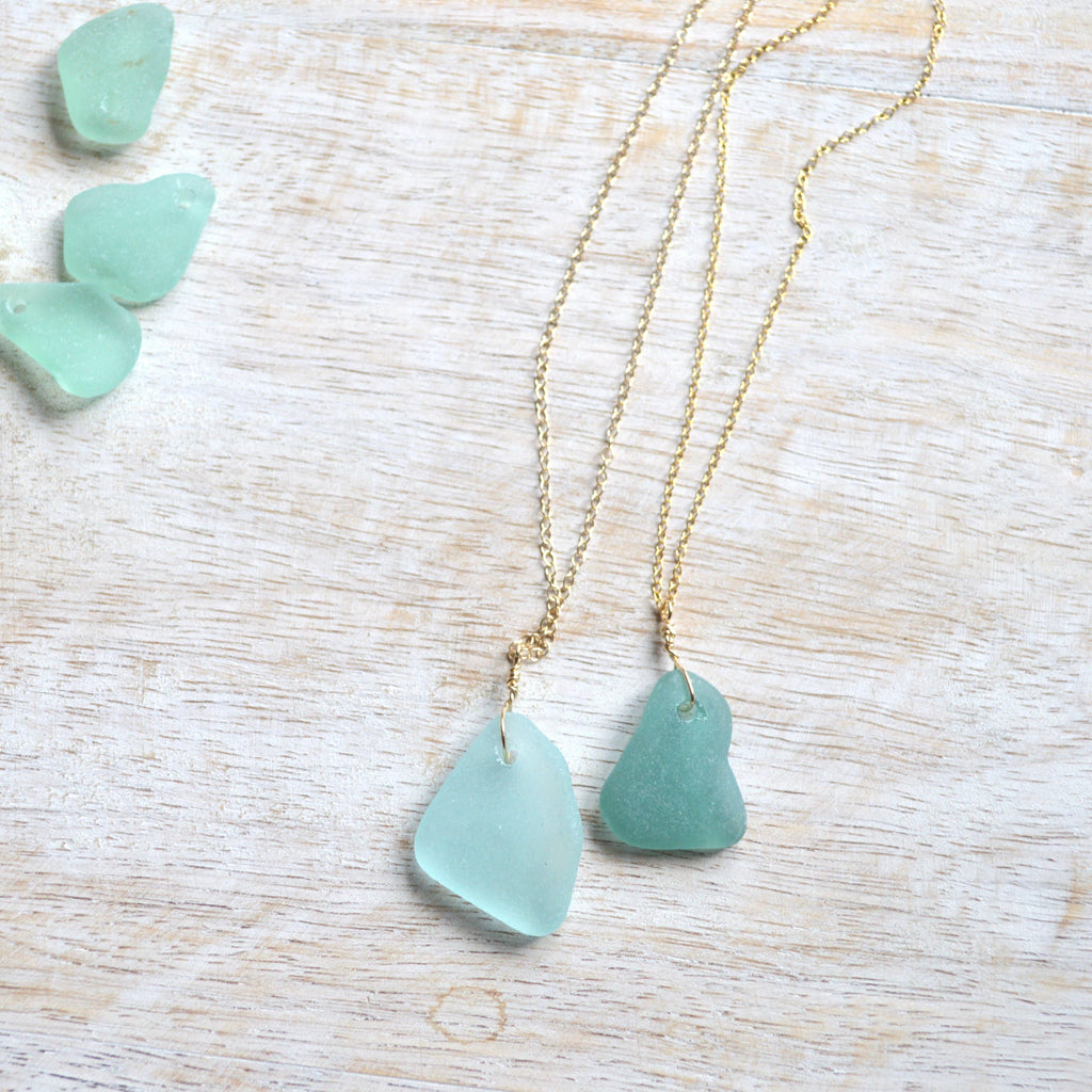 Two gold chain necklaces with irregular light green sea glass pendants, displayed on a light wood surface with three matching sea glass chips in the upper left corner.