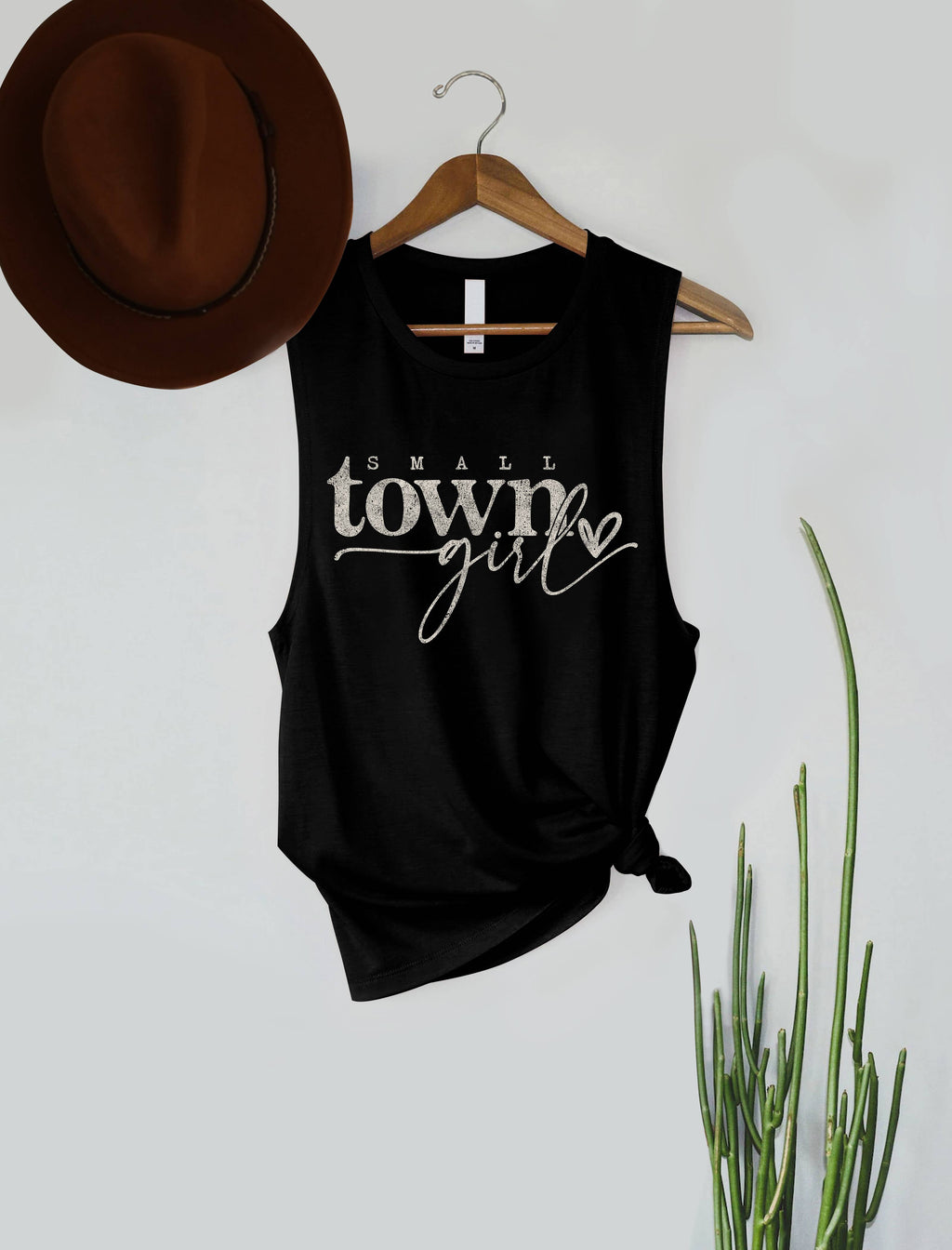 A black sleeveless tank top with a front knot and white 'Small town girl' text, hanging on a wooden hanger against a light gray wall. A brown hat is partly visible at the top left, and green plant stems appear at the bottom right.