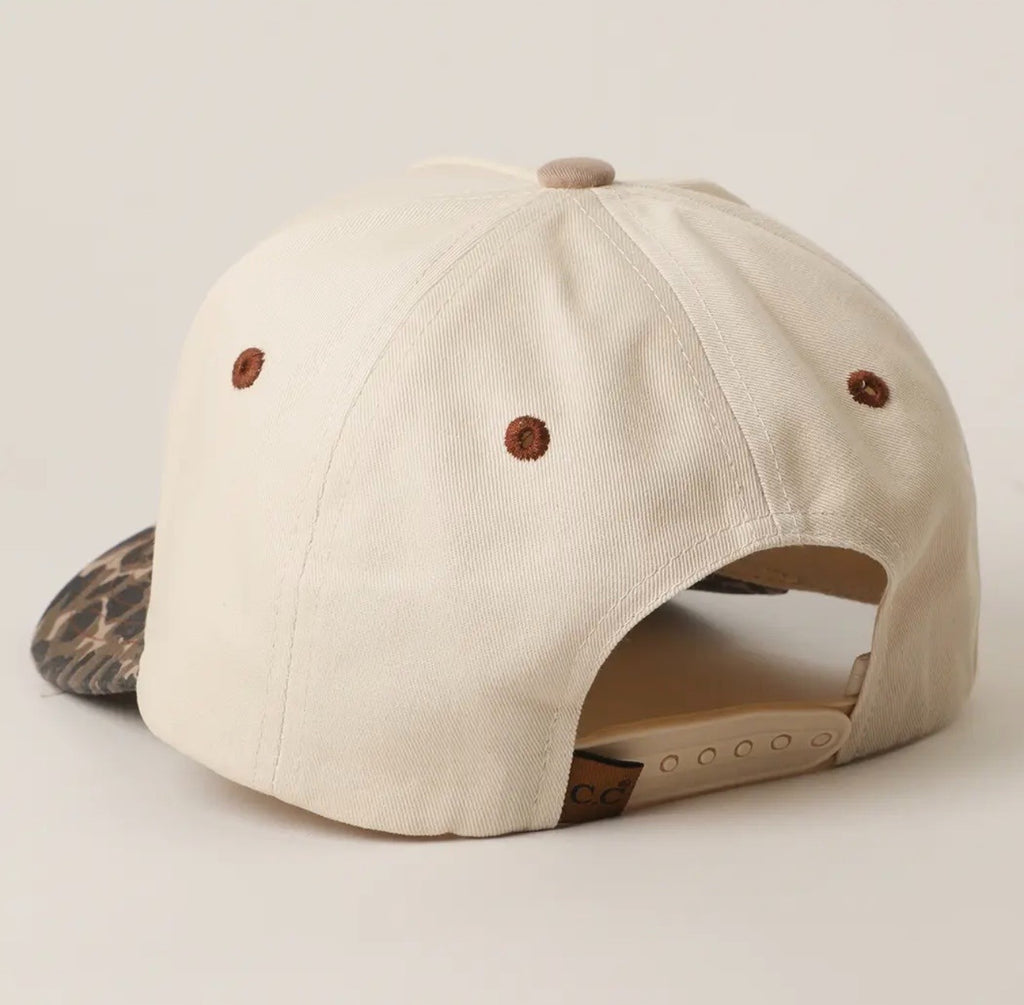 An off-white canvas baseball cap shown from a rear three-quarter angle, featuring brown embroidered eyelets, a tan adjustable strap, and a patterned visor peeking out on the left.