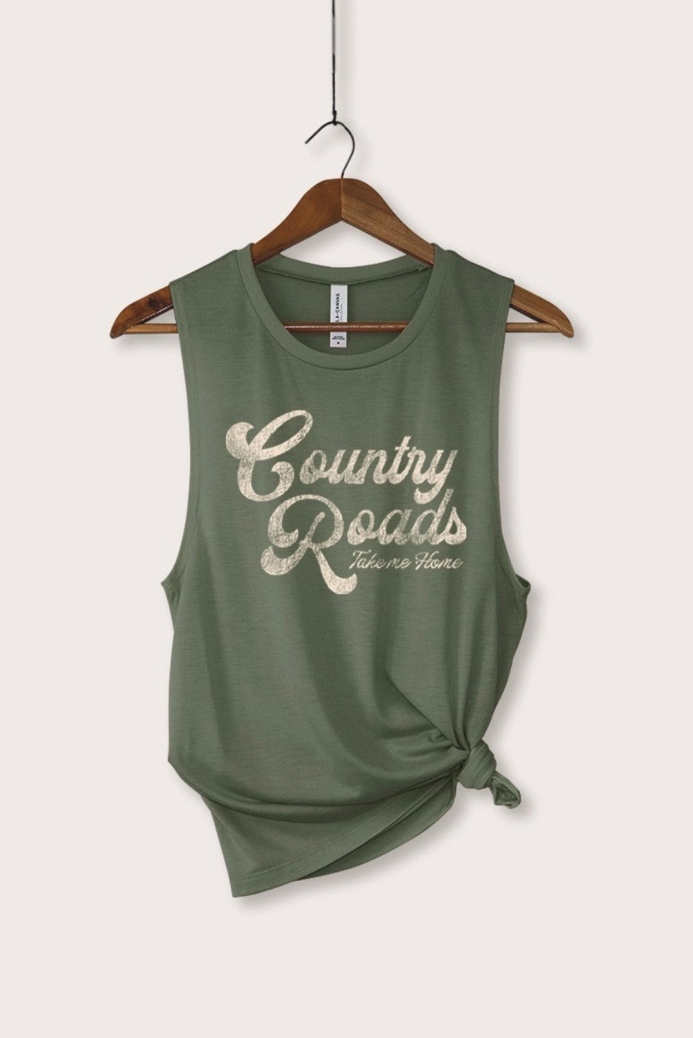 An olive green sleeveless tank top with a distressed cream graphic reading 'Country Roads Take Me Home', hanging on a wooden hanger against a light beige background.