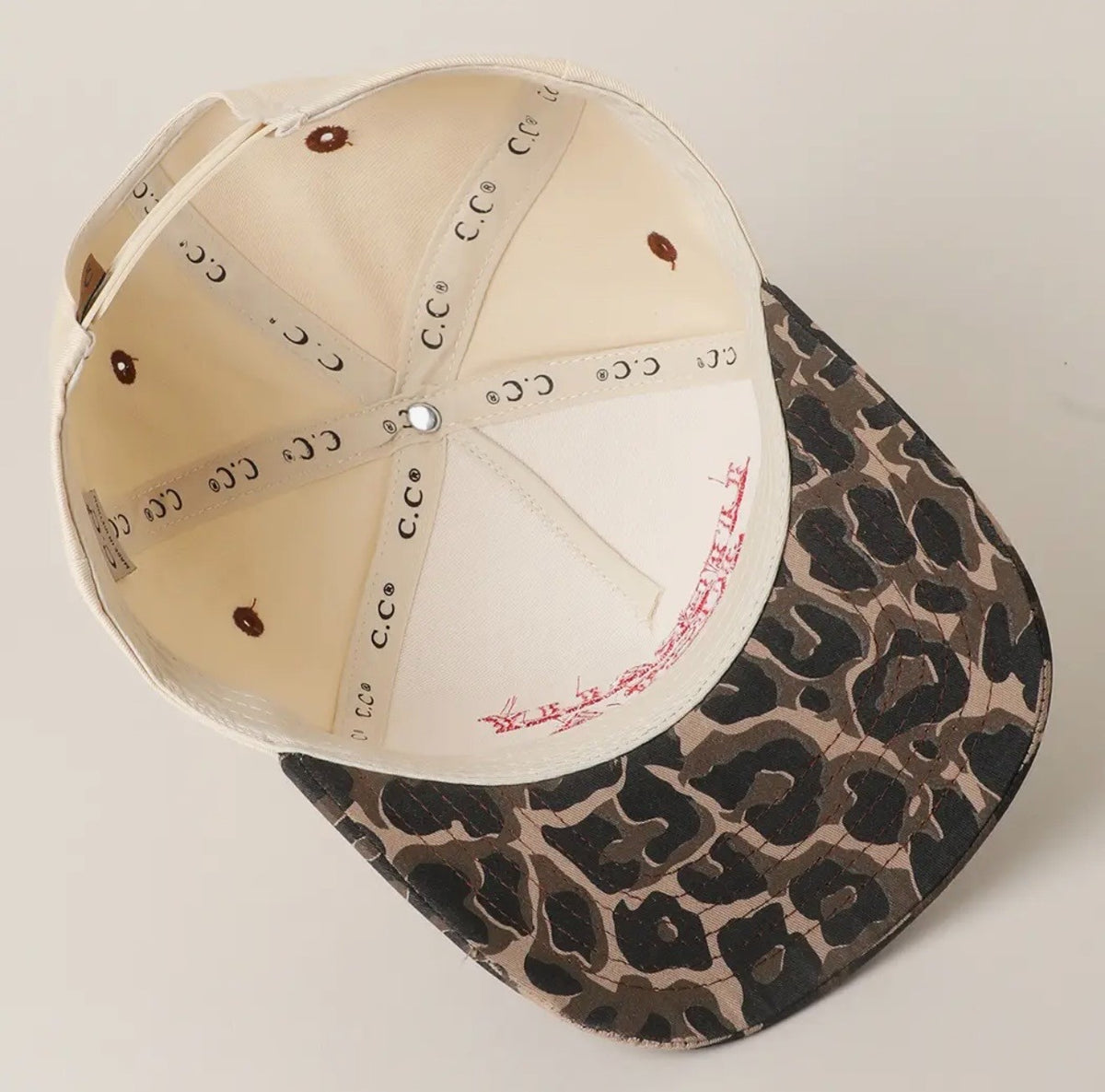 A structured baseball cap shown from an overhead angle, featuring a cream-colored interior with printed elastic bands and a leopard-print patterned exterior in brown, black, and beige.