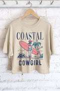 Beige t-shirt with 'Coastal Cowgirl' graphic design on a hanger against a white brick wall.