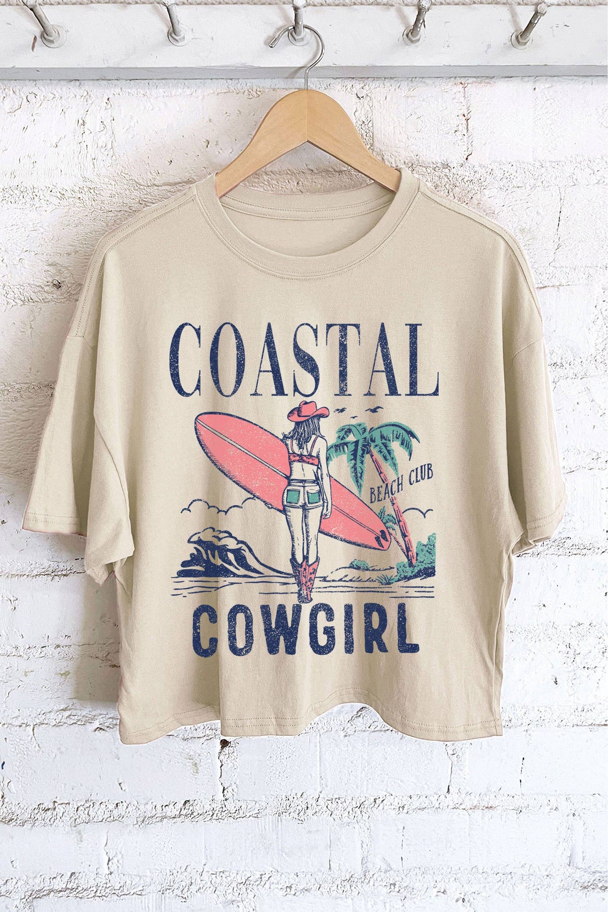 Beige t-shirt with 'Coastal Cowgirl' graphic design on a hanger against a white brick wall.