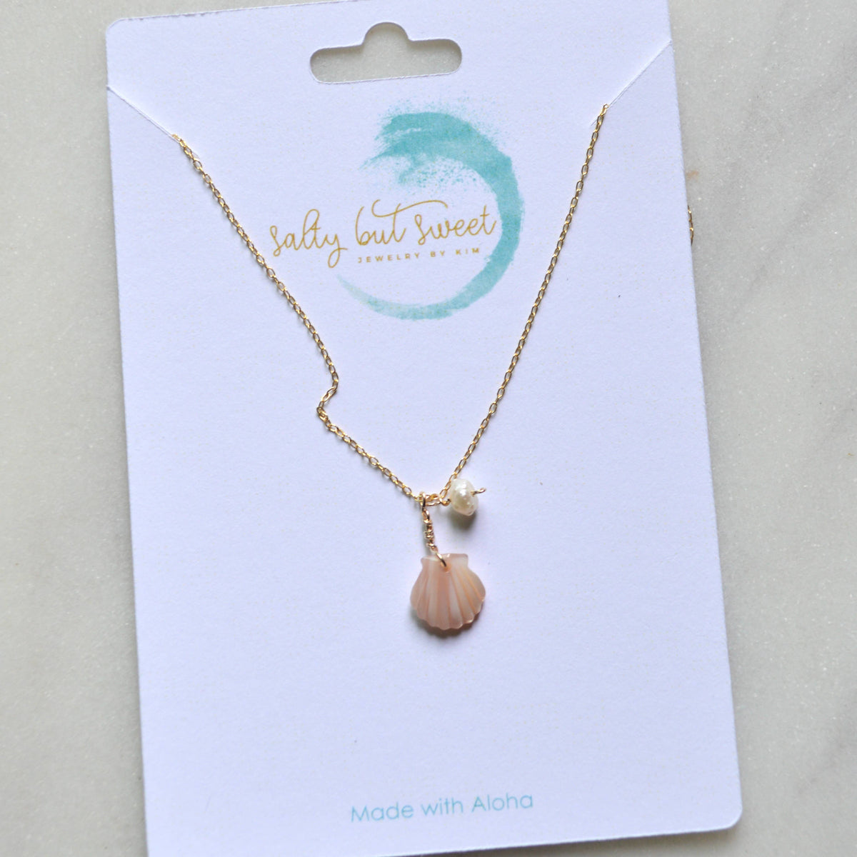 A gold chain necklace with a small peach-pink seashell pendant and a tiny white pearl-like bead, displayed on a white jewelry card branded 'sally but sweet'.