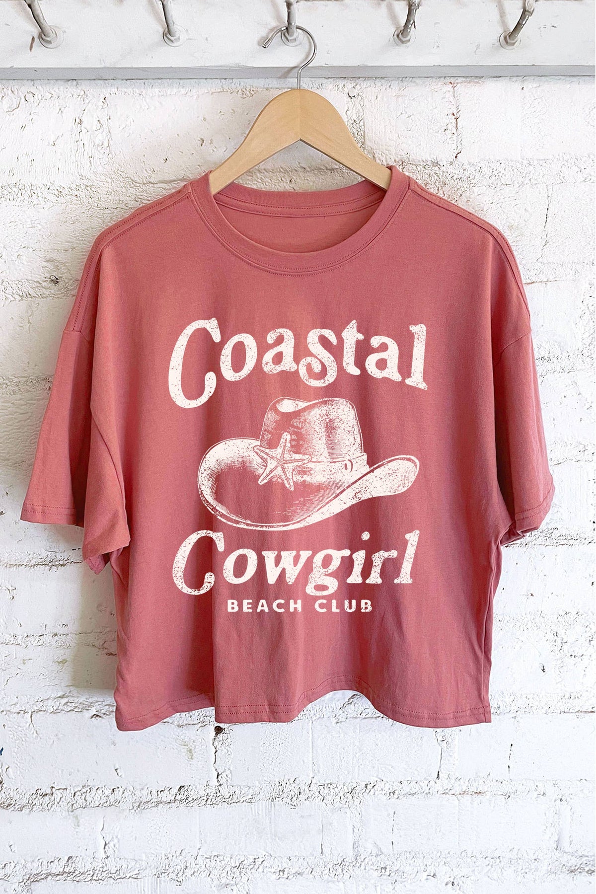 Pink t-shirt with 'Coastal Cowgirl Beach Club' text and graphic on a hanger against a white brick wall.