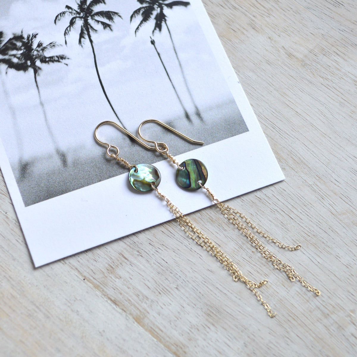 A pair of gold-tone dangle earrings with round iridescent abalone shell beads and long chain drops, displayed on a white card over a light wood surface.