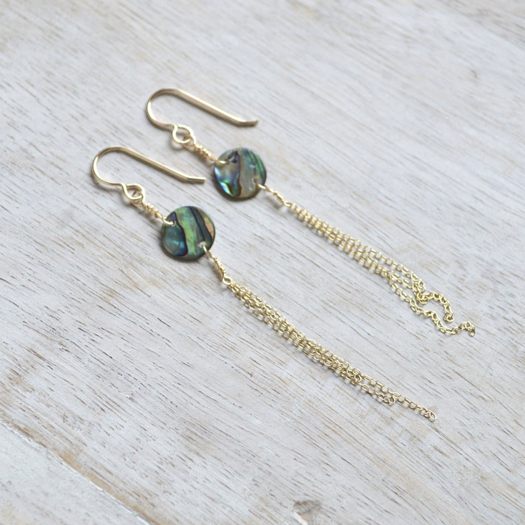 A pair of gold-tone dangle earrings with round iridescent abalone shell beads and long chain drops, displayed on a light wood surface.