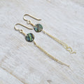 A pair of gold-tone dangle earrings with round iridescent abalone shell beads and long chain drops, displayed on a light wood surface.