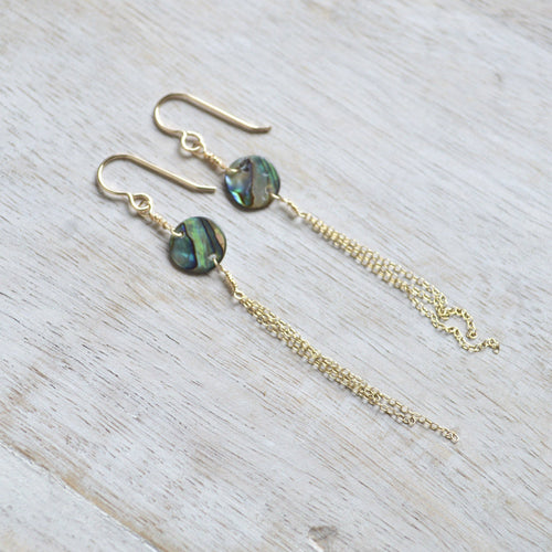A pair of gold-tone dangle earrings with round iridescent abalone shell beads and long chain drops, displayed on a light wood surface.