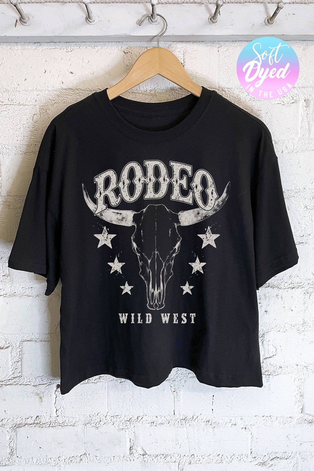 A black short-sleeve graphic t-shirt hanging on a wooden hanger against a white painted brick wall. The shirt features a distressed white graphic of a bull skull with 'RODEO' above and 'WILD WEST' below, accented with stars.