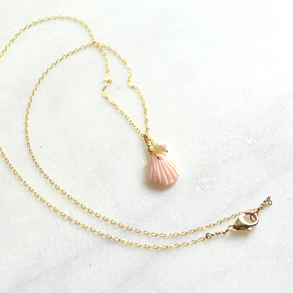 A delicate gold chain necklace featuring a small, realistic pink seashell pendant, displayed on a light, textured white surface.
