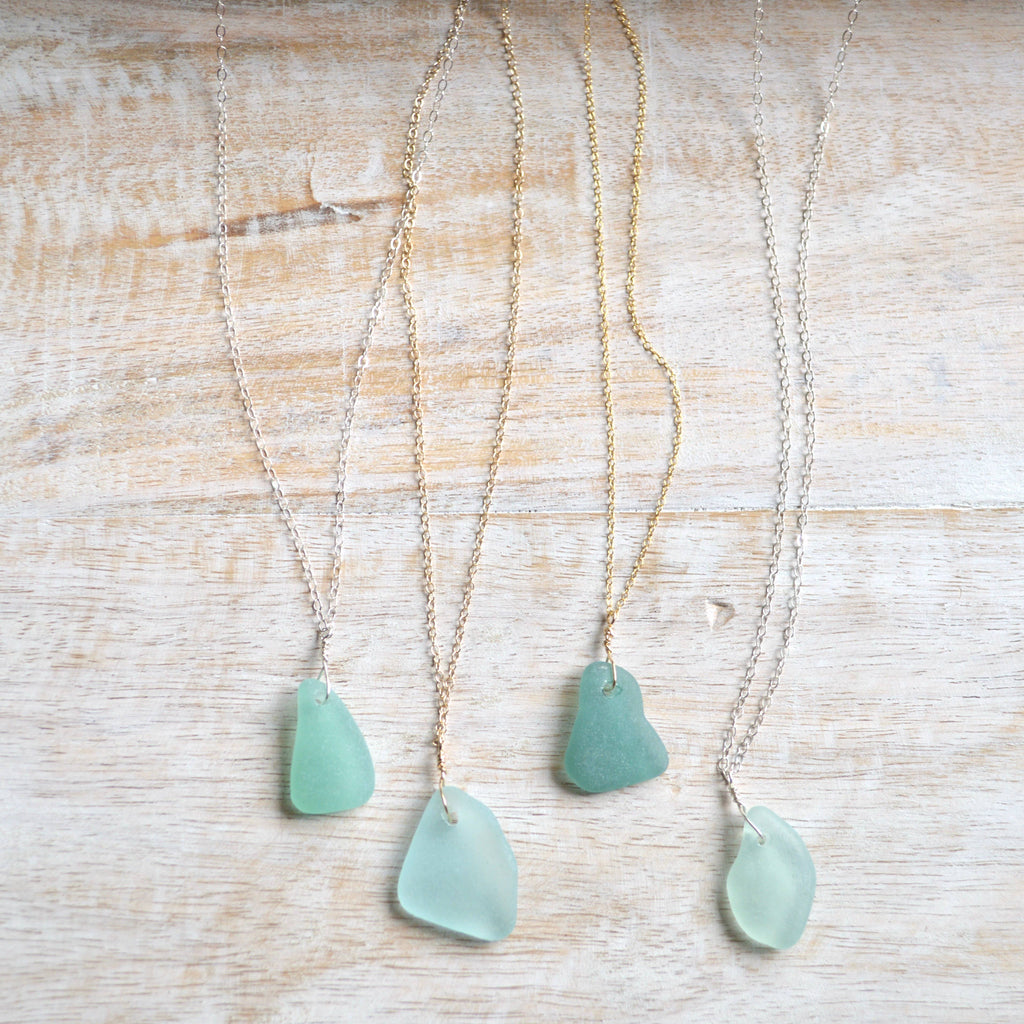 Four sea glass pendants on delicate chains, displayed on a light wood background. The pendants are in various shades of seafoam green and light blue-green, paired with both gold and silver chains.