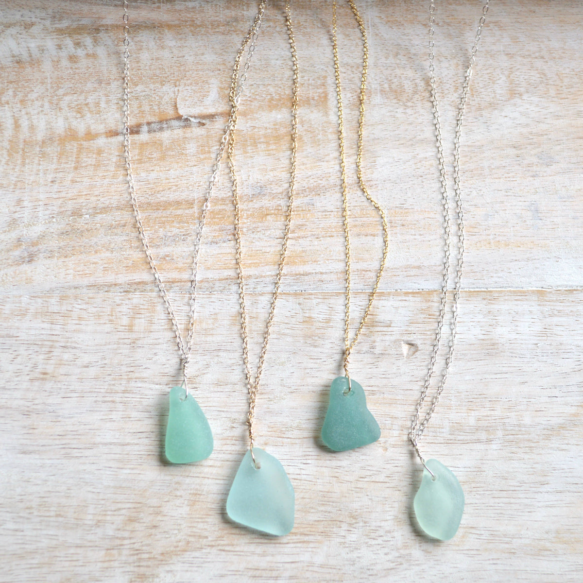 Four sea glass pendants on delicate chains, displayed on a light wood background. The pendants are in various shades of seafoam green and light blue-green, paired with both gold and silver chains.