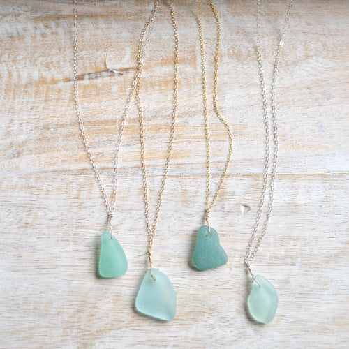 Four sea glass pendants on delicate chains, displayed on a light wood background. The pendants are in various shades of seafoam green and light blue-green, paired with both gold and silver chains.
