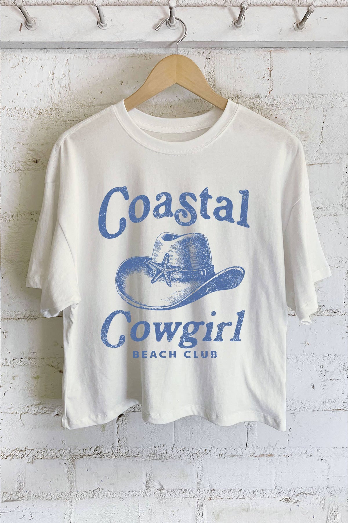 White t-shirt with 'Coastal Cowgirl Beach Club' text and graphic on a hanger against a white brick wall.